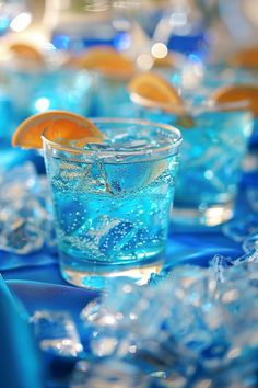 there are many glasses with blue liquid and orange slices on the rims that have ice cubes around them