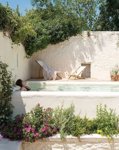 an outdoor hot tub surrounded by flowers and greenery next to a wall with a woman sitting in it