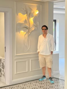 a man standing in front of a white wall with flowers on it and lights behind him