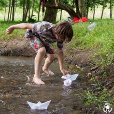 a little boy playing with paper boats in the water near some trees and grass,