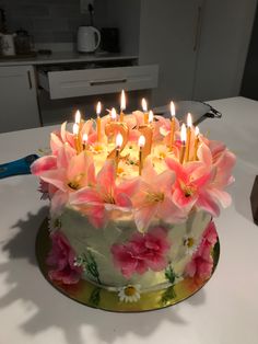 a birthday cake with pink flowers on it and lit candles in the middle, sitting on a kitchen counter