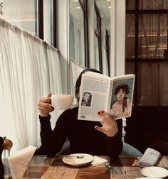 a woman sitting at a table reading a book and holding a coffee cup in front of her face