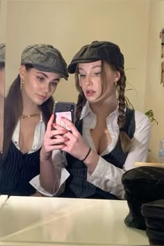 two women are looking at their cell phones in front of the mirror, one is wearing a hat