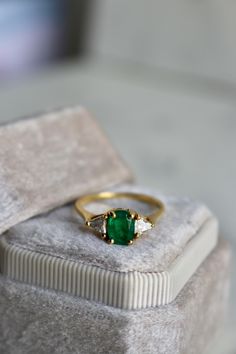 One Vintage Emerald Diamond 18k Yellow Gold Ring. Featuring one cushion cut emerald weighing approximately 0.70 carat. Accented by two trillion cut diamonds with a total weight of approximately 0.30 carat, graded F color, VS clarity. Crafted in 18 karat yellow gold with purity mark. Circa 1980. The ring is a size 6 ½ and may be resized.About this Item: This ring dazzles with timeless elegance. A cushion cut emerald of approximately 0.70 carat shines at the center, evoking the glamour of decades past. Two trillion cut diamonds totaling 0.30 carat twinkle on either side, adding subtle sparkle that catches the light. The emerald's vivid hue and diamonds' fiery fire combine to create a color symphony on your finger. Crafted from 18 karat yellow gold, this ring is designed to complement a varie Engagement Ring Quiz, Colored Stone Engagement Rings, Halo Style Engagement Rings, Retro Era, Jewelry Education, Colored Engagement Rings, 18k Yellow Gold Ring, Stone Engagement Rings, Shop Engagement Rings