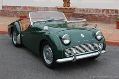 an old green convertible car parked in front of a fountain with potted planters