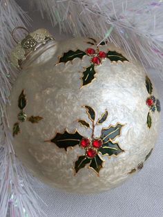 a white christmas ornament with holly leaves and red berries hanging from a tree