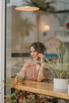 a woman talking on her cell phone while sitting at a table in front of a window