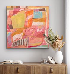 an abstract painting hangs on the wall next to a wooden cabinet and vase with flowers