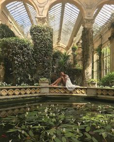 a woman sitting on the edge of a pond surrounded by plants and greenery in a greenhouse