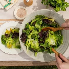 two hands holding wooden spoons over a bowl of lettuce on a picnic table