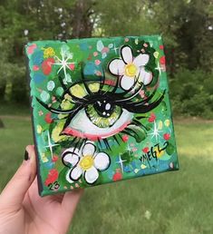 a hand holding up a small square piece of art with flowers and eyes painted on it