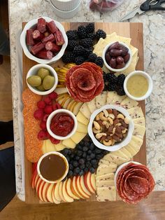 an assortment of cheeses, meats and fruit on a cutting board with dipping sauces