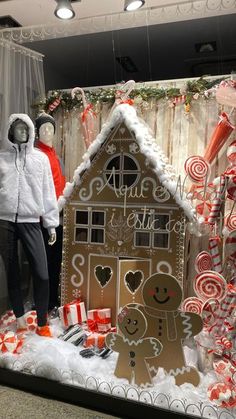 two mannequins dressed in winter clothing stand next to a gingerbread house