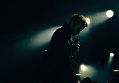 a man holding a microphone in front of some spotlights on a dark stage with lights behind him