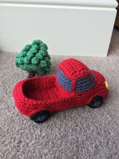 a crocheted red car next to a small green tree on the floor in front of a white wall