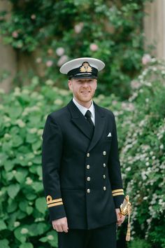 a man in a navy uniform standing next to some bushes and flowers, smiling at the camera