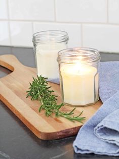 a wooden cutting board topped with a candle next to a glass jar filled with greenery