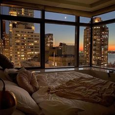 an unmade bed in front of a large window overlooking the city at night time