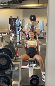 a woman is sitting on a bench in the gym while a man looks at her phone