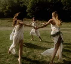 two women in white dresses holding hands and running across the grass with trees in the background