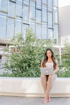 a woman sitting on a ledge with a laptop in her lap and looking at the camera