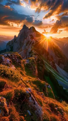 the sun is setting on top of a mountain with grass and rocks in the foreground