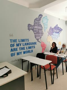 two women sitting at desks in front of a wall with words written on it