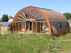 a house made out of wood and glass in the grass with a large wheel on it's side