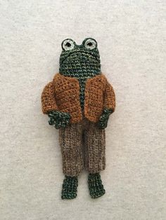 a crocheted frog wearing a sweater and pants with eyes wide open, standing on a white surface