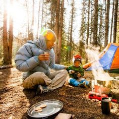 a man and child sitting on the ground near a tent in the woods with food