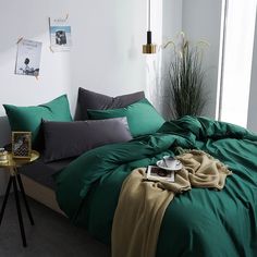 a bed with green sheets and pillows in a white room next to a plant on a table