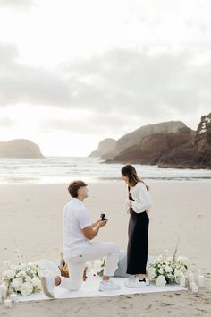 a man kneeling down next to a woman on top of a white blanket at the beach