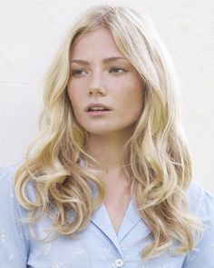 a woman with long blonde hair standing in front of a white wall wearing a blue shirt