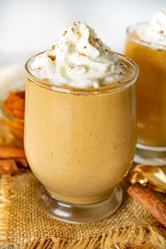 two glasses filled with whipped cream on top of a cloth covered place mat next to cinnamon sticks