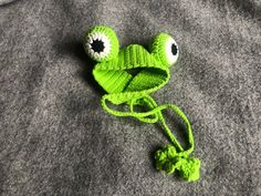 a crocheted frog hat with eyeballs on it's face laying on a gray surface