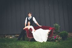 a bride and groom sitting on a red couch in front of a barn wall with flowers