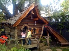 two children are standing in front of a small house made out of logs and wood