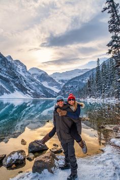 two people standing on the shore of a lake with mountains in the background