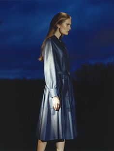 a woman in a blue leather coat is standing on a dark background with her hands behind her back