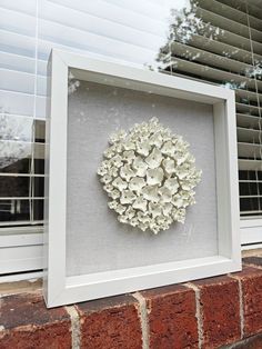 a white framed artwork with flowers in it on a brick wall next to a window