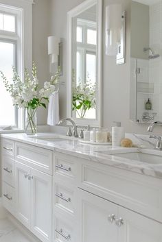 a white bathroom with double sinks and large mirrors on the wall, along with two vases filled with flowers