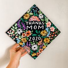 a hand holding a graduation cap that says thanks mom on it with flowers and leaves