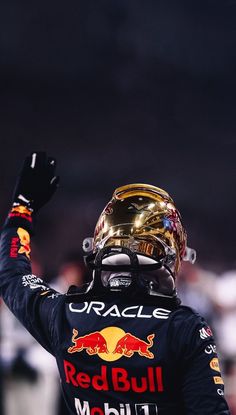 a man in a red bull racing uniform raising his fist up to the sky while wearing a gold helmet