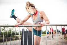 a woman leaning on a fence with her leg up and looking at the ground while other people are running in the background