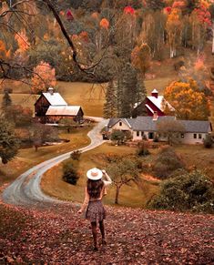 a woman standing in the middle of a field with autumn leaves on the ground and houses behind her