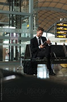 a man in a suit sitting on an airport bench looking at his tablet