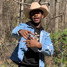 a young man wearing a cowboy hat and denim jacket standing in front of some trees