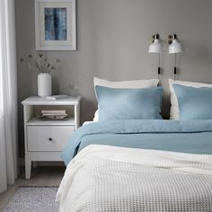 a bed with blue sheets and pillows in a white room next to a window on a wooden floor