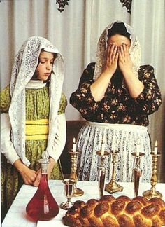 two women dressed in medieval clothing stand at a table with bread and candles on it