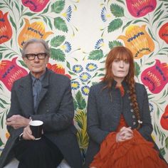 two people sitting next to each other in front of a wall with flowers on it